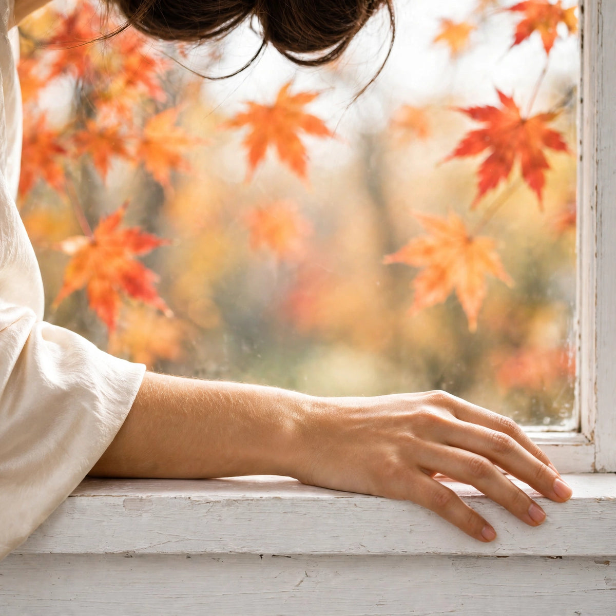 Hand on a white windowsill with autumn maple leaves outside — YNA Parfums handcrafted in Canada